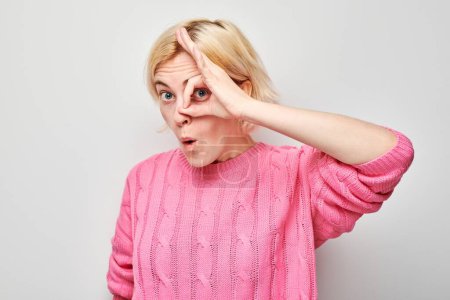 Téléchargez les photos : Femme gaie en pull rose faisant signe OK avec la main sur les yeux, souriant sur fond clair. - en image libre de droit