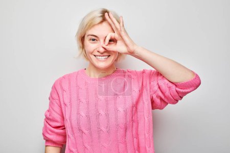 Cheerful woman in pink sweater making OK sign with hand over eye, smiling on light background.