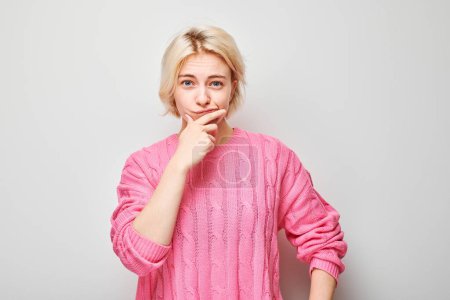 Nachdenkliche junge Frau im rosafarbenen Pullover mit der Hand am Kinn, die in die Kamera auf hellgrauem Hintergrund blickt.