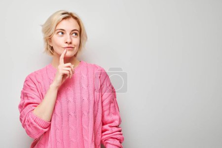 Young woman in pink sweater looking thoughtful with finger on cheek, isolated on light background.