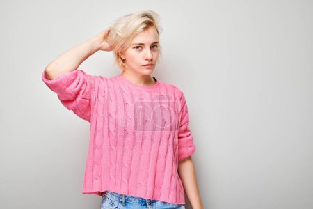 Téléchargez les photos : Portrait d'une jeune femme dans un pull rose posant avec désinvolture avec une main dans les cheveux sur fond gris. - en image libre de droit