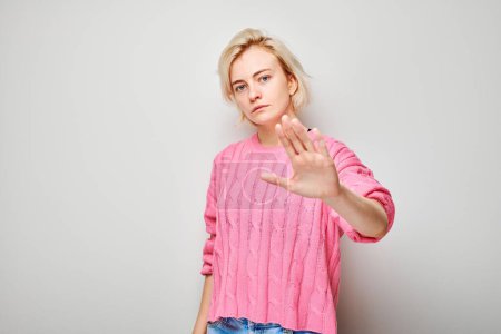 Téléchargez les photos : Jeune femme confiante en pull rose montrant geste d'arrêt avec la main sur fond clair. - en image libre de droit