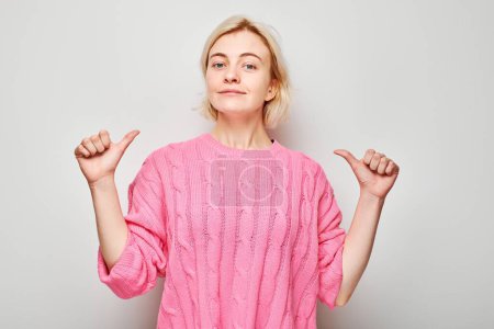 Photo for Confident woman in pink sweater pointing at herself with thumbs, isolated on white background. - Royalty Free Image