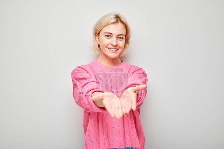 Téléchargez les photos : Femme souriante en pull rose tenant les mains dans un geste accueillant sur un fond gris. - en image libre de droit