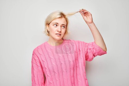 Verwirrte junge Frau im rosafarbenen Pullover blickt auf ihr Haar vor weißem Hintergrund.
