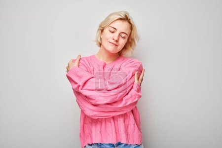 Photo for Portrait of a content woman in a pink sweater hugging herself against a gray background. - Royalty Free Image