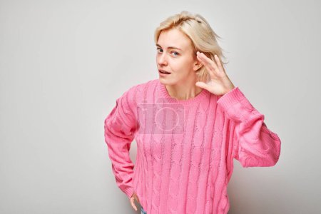 Téléchargez les photos : Femme en pull rose écoutant avec la main à l'oreille, fond blanc. - en image libre de droit