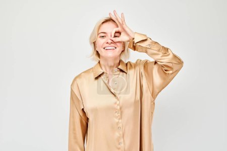 Téléchargez les photos : Femme souriante en chemisier beige faisant OK geste au-dessus des yeux, isolé sur fond clair. - en image libre de droit