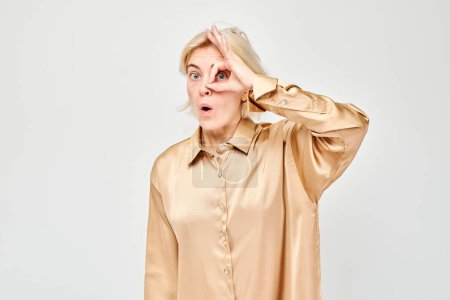 Foto de Mujer sorprendida en blusa beige haciendo buen gesto sobre el ojo, fondo blanco. - Imagen libre de derechos