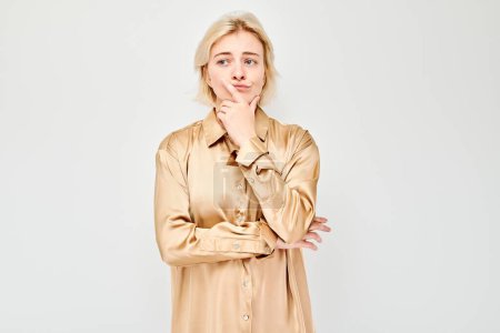 Téléchargez les photos : Jeune femme réfléchie en chemisier beige avec la main sur le menton à la recherche coûteuse sur un fond blanc. - en image libre de droit