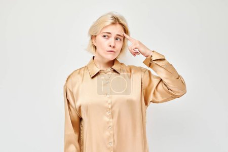 Téléchargez les photos : Jeune femme confus en chemisier beige geste avec le doigt à la tête sur un fond gris. - en image libre de droit