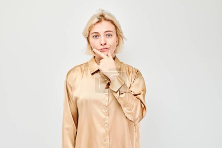 Thoughtful young woman in a beige blouse, hand on chin, looking pensive against a white background.