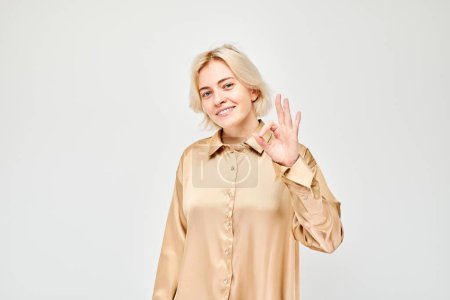 Photo for Smiling woman in a beige blouse giving an OK hand sign against a light background. - Royalty Free Image