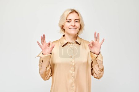Photo for Smiling woman in beige shirt making OK sign with both hands on a white background. - Royalty Free Image