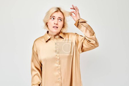 Verwirrte junge Frau in beiger Bluse hält Haarsträhne mit trockenen Enden isoliert auf grauem Hintergrund