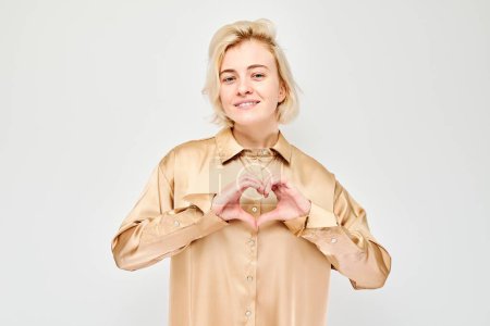 Téléchargez les photos : Femme souriante en forme de coeur avec les mains, portant une chemise beige sur fond blanc. - en image libre de droit