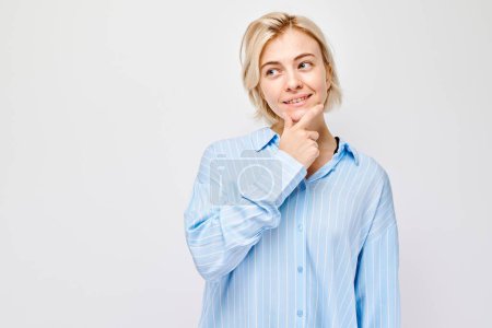 Téléchargez les photos : Femme réfléchie avec un sourire ludique touchant menton, portant une chemise bleue sur un fond blanc. - en image libre de droit