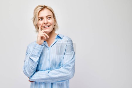 Téléchargez les photos : Portrait de femme réfléchie souriant avec la main sur le menton en chemise bleue sur fond gris clair. - en image libre de droit