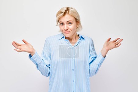 Foto de Mujer de camisa azul encogiéndose de hombros con expresión incierta y juguetona sobre un fondo claro. - Imagen libre de derechos