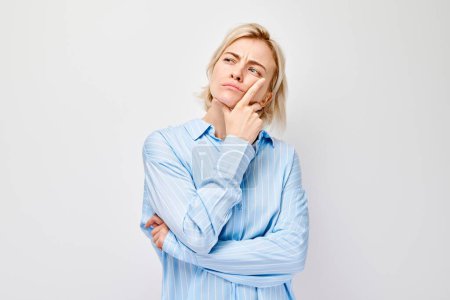 Téléchargez les photos : Jeune femme réfléchie avec la main sur le menton, portant une chemise bleue, isolée sur un fond blanc. - en image libre de droit