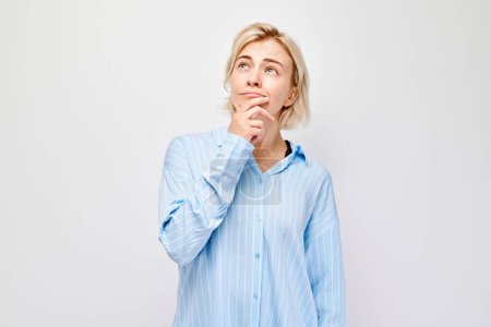 Téléchargez les photos : Jeune femme réfléchie avec la main sur le menton, portant une chemise bleue, isolée sur un fond blanc. - en image libre de droit