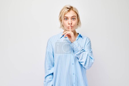 Foto de Retrato mujer en camisa azul, haciendo gesto de silencio con el dedo en los labios sobre fondo blanco. - Imagen libre de derechos