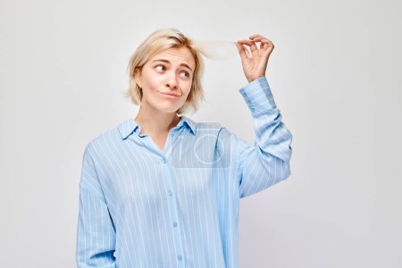 Photo for Woman in blue shirt looking puzzled, twirling hair, white background. - Royalty Free Image