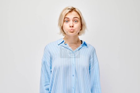 Téléchargez les photos : Jeune femme confus en chemise bleue haussant les épaules avec des joues gonflées sur un fond gris. - en image libre de droit