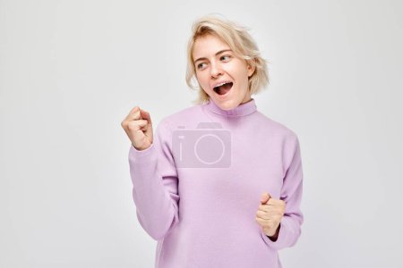 Portrait of smiling face blond young woman clenching fists and rejoicing, celebrating victory isolated on white studio background, advertising banne