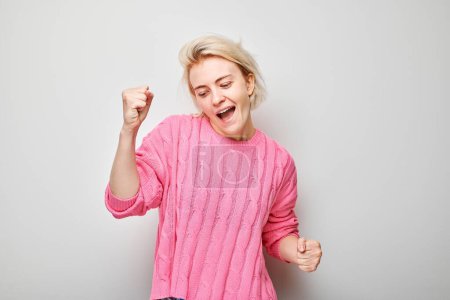 Portrait of smiling face blond young woman clenching fists and rejoicing, celebrating victory isolated on white studio background, advertising banne