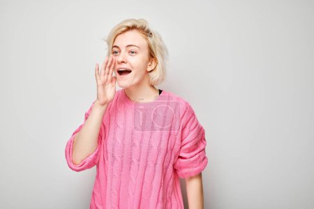 Téléchargez les photos : Portrait de fille blonde criant bruyamment avec les mains, nouvelles, paumes pliées comme un mégaphone isolé sur fond blanc - en image libre de droit