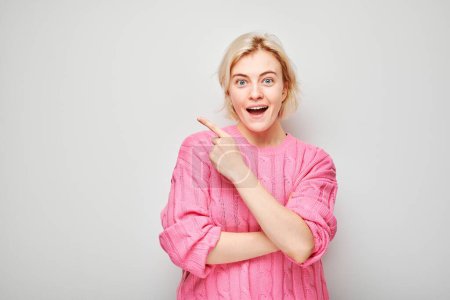 Téléchargez les photos : Jeune femme blonde en pull rose pointant du doigt, démontrant un espace vide pour le produit ou le texte isolé sur fond de studio blanc - en image libre de droit