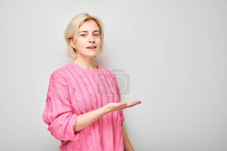Photo for Young blond woman in pink sweater holding something in hand, demonstrating empty space for product or text isolated on white studio backgroun - Royalty Free Image
