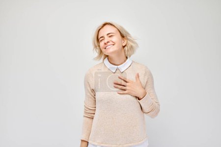 Photo for Happy young woman smiling with eyes closed, hand on heart, expressing gratitude or contentment, isolated on a light background. - Royalty Free Image