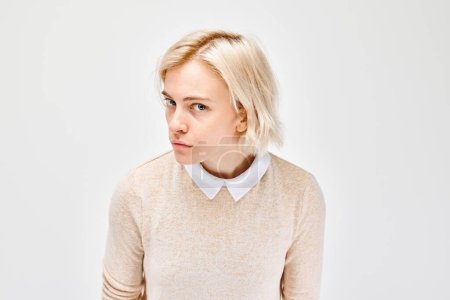 Photo for Skeptical young woman with short blonde hair, wearing a beige sweater over a collared shirt, glancing sideways against a white background. - Royalty Free Image