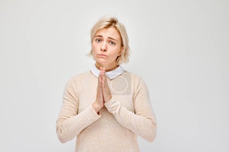 Adult woman with hopeful expression, hands pressed together in pleading gesture, isolated on light background.