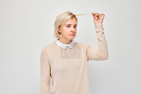 Portrait of blond woman holding her lock of hair, dissatisfied with quality of hair, dry and split ends
