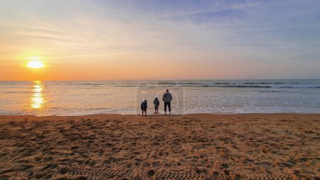 Foto de Padre con dos hijos se quedan en la playa de arena cerca del mar. Fondo de puesta de sol. - Imagen libre de derechos