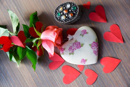 Corazón de porcelana con estampado de flores en la mesa textural de madera, rosa rosa y muchos corazones de papel rojo alrededor. Acostado. Regalo y saludo para el Día de San Valentín o el Día de las Madres. 
