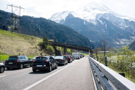 A lot of cars stay in traffic jam on the road near tunnel with mountains and forest in Switzerland.