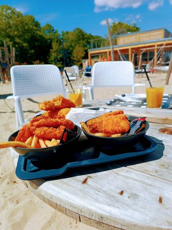 Fastfood on the table on the beach fries and fried chicken fish in bowls with ketchup beach chairs. High quality photo. Juice, white sand. Rest on the beach in the cafe with street food. Family time.