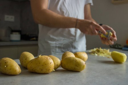 Téléchargez les photos : Un homme épluchant des pommes de terre sur un comptoir de cuisine gris avec un éplucheur de pommes de terre - en image libre de droit