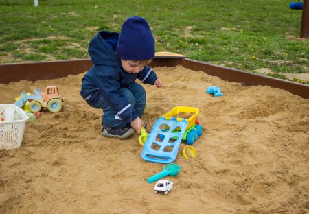 Toddler boy, playing outdoors in a sandbox with a green toy trowel and blue car. High quality photo