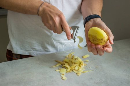 Téléchargez les photos : Un homme épluchant des pommes de terre sur un comptoir de cuisine gris avec un éplucheur de pommes de terre. Photo de haute qualité - en image libre de droit