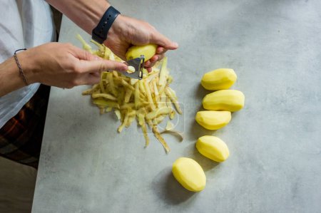 Téléchargez les photos : Un homme épluchant des pommes de terre sur un comptoir de cuisine gris avec un éplucheur de pommes de terre. Photo de haute qualité - en image libre de droit