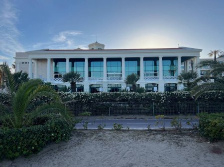 Esta imagen muestra un elegante hotel frente al mar rodeado de exuberantes palmeras, situado en un impresionante telón de fondo al atardecer. La arquitectura de los hoteles emana sofisticación y lujo, complementado por la