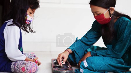 Two girls wearing masks play Dakon, a traditional Indonesian children's game