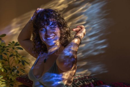 Foto de Portrait of a smiling young woman with curly dark hair lifting her arms in the air as if dancing in refracted light - Imagen libre de derechos
