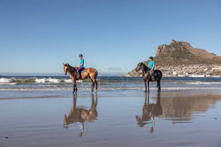 Two teenage girls on horseback cantering in the water at low tide on the beach under a cloud sky with a beautiful mountain in the background