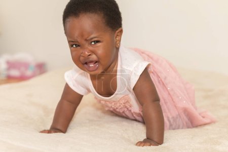 Portrait of a crying black baby girl dressed in a pink dress crawling on a bed